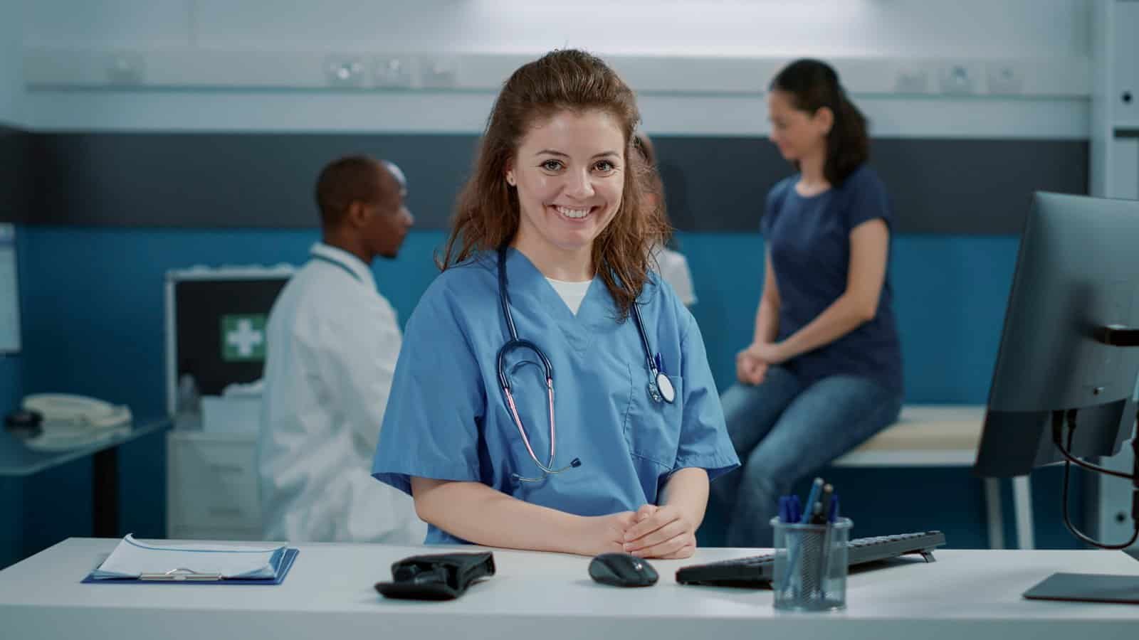 portrait woman nurse smiling wearing uniform office sitting desk medical assistant with stethoscope looking camera getting ready help doctor appointment scaled