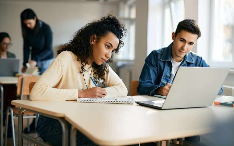 High school classmates taking notes while using laptop in the classroom.